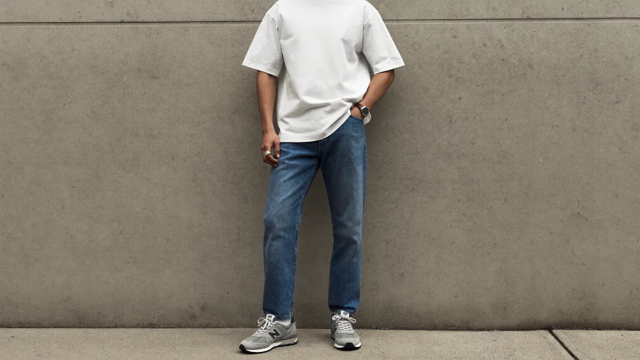 Homme en t-shirt blanc, jean droit et sneakers grises devant un mur de béton